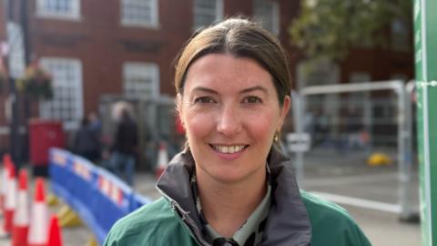 A woman - Sarah Walmsley - smiling straight at the camera. She is wearing a green coat and is standing in front of a set of roadworks.