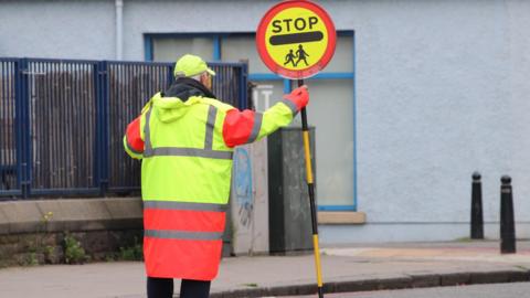 A lollipop man wearing a hi-vis jacket standing in the middle of a road holding a stop sign.