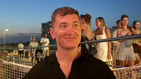 Will Bailey looks to the side as he smiles. He is sitting down wearing a black shirt. People can be seen walking on a walkway behind him.