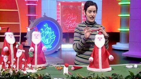 Romana D'Annunzio stands behind a green table, making a Santa table decoration. On her right are a group of completed Santa decorations