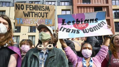 A group of protesters hold up placards. One reads "No feminism without trans women". Another reads "lesbians love trans women" and is being held up by a woman wearing a pink cardigan and a pink face mask. 