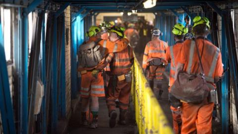 Coal miners wearing orange overalls, hardhats and protective equipment hug and engages with each other as they walk through a corridor after finishing their final shift before closure at the Kellingley Colliery in Yorkshire.