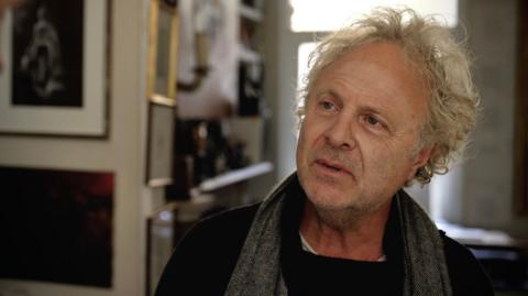 Charlie Mackesy with a scarf around his neck, in his home with pictures on the wall, he is highlighted by sun from the window behind him
