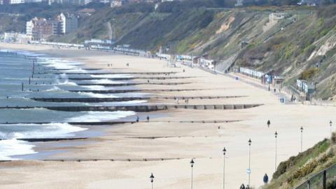 A shot from a beach taken from higher ground. Waves can be seen lapping the shore and the small figures of a few people can be seen on the sand.