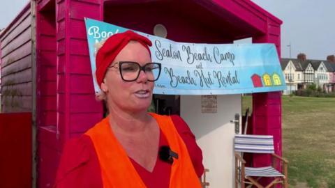 Woman wearing a high viz jacket, large spectacles and a red bandana standing in front of a beach hut with a banner reading "Seaton Beach Camp and Beach Hut Rental.