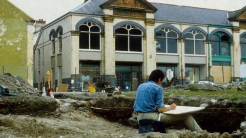 An old picture of a man at an archaeological excavation site. He is sat down with a board on his lap. There is a building in the background with mounds of dirt around him. 