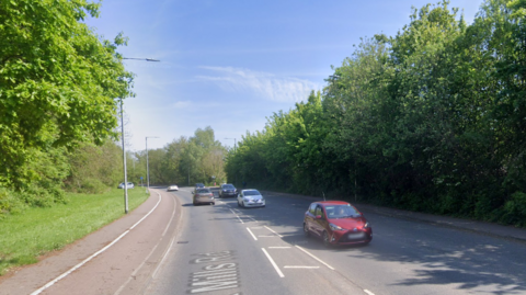 A grey tarmac road with traffic on both sides. A red car is in the foreground coming towards the camera. The road is flanked by bushes and trees.