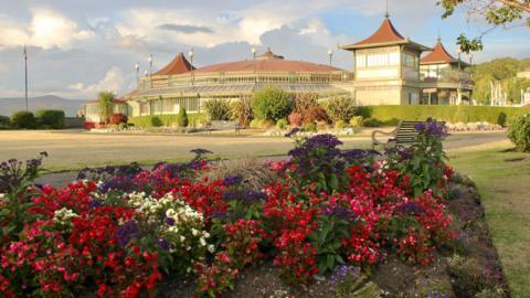 A park with a Victorian-era pavilion in the background. Colorful flowerbeds are in the foreground, and the sky is filled with dramatic clouds.