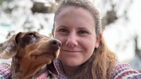 Molly Leonard with a tan and brown Dachshund. Molly is smiling at the camera wearing a pink, purple and yellow knitted jumper, her long brown hair is tied back.