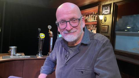 A man with a bald head, glasses and a white beard sits in a bar on a theatre stage. He is wearing a grey t-shirt under a blue-grey shirt.