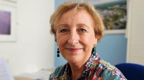 Prof Lesley Dwyer, who has short hair and a floral top, sitting in her office and smiling