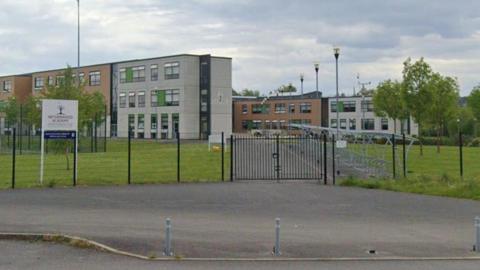 A street view image of the school gate. It shows a modern multi-storey complex with several smaller buildings in either white or brown attached. The main buildings sit in front of a big lawn which has a row of cycle storage to the left.