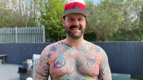Tom Radford stands bare chested showing his tattoos. He has a black beard and moustache and is wearing a red Spiderman baseball cap