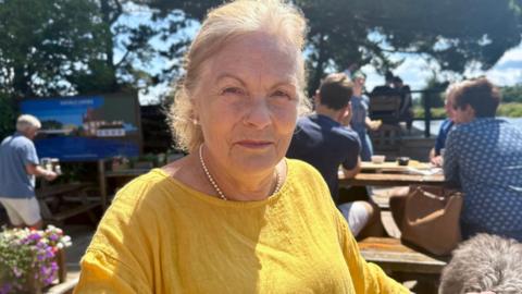 Annie Lewis Marffy in a yellow top sitting outdoors at a wooden table. In the background, there are other people seated at tables, trees, and a blue sky with some clouds. 

