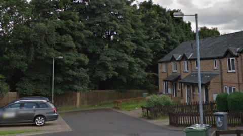 A residential street with cars parked on one side and a number of terraced homes on the other.