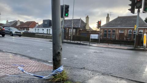 Road with cars on it with houses down either side. There is a pedestrian crossing with police tape attached to it.