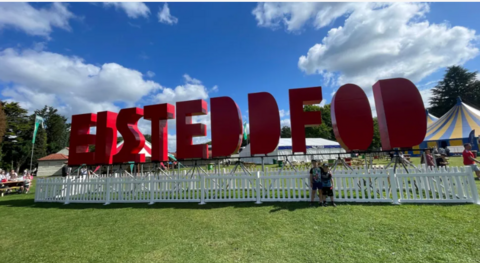 Big red letters spelling out Eisteddfod with th stalls and tents of the event in the background and green grass in front