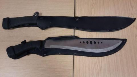 Two large zombie knives with black cases. The knives have seven holes in the middle and black handles.