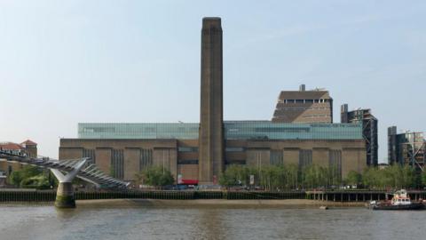 The exterior of London's Tate Modern gallery, viewed from across the river. The River Thames is visible in the foreground at the bottom of the picture, with a grey pedestrian bridge spanning the river in the bottom left corner. A large brown building is situated on the river bank, with a top floor made of glass. A large brown tower rises from the front-middle of the building, by the riverside. There is a row of green leafy trees along the river bank. The sky is pale blue and sunny.