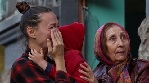 A young woman cries with her hand over her face as she holds a small child. A second, older woman wearing a scarf over her head puts her hand on the child's shoulder. 