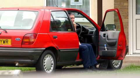 Huntley is half-sitting in a red Fiesta car, parked outside a house with a white front door. He is wearing dark blue jeans or trousers. He is sitting sideways in the driver's seat. The driver's side door is open and he has his feet on the ground.