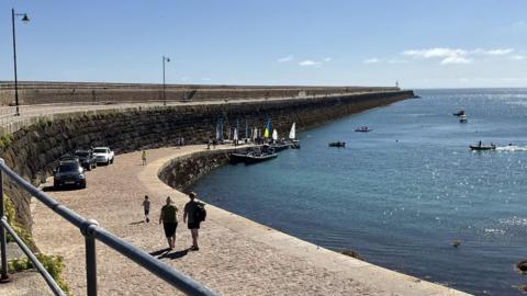 St Catherine's harbour. This view shows a sea wall on the left sweep around to the right. There are people and cars on the breakwater. Some small yachts can be seen in full sail on a slipway and in the sea on what is a sunny day. 