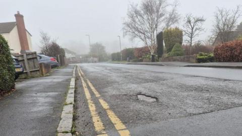 A badly-surfaced road, with a large pothole near double-yellow lines. The road is quite wide and runs though a residential area, with pavement either side and houses, cars, driveways and street furniture like streetlamps visible. The picture has been taken on a foggy day. 