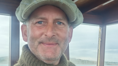 Jeremy Gower grins at the camera in the wheelhouse of his boat. He is wearing a large grey woollen cap and a khaki green roll-neck jumper.