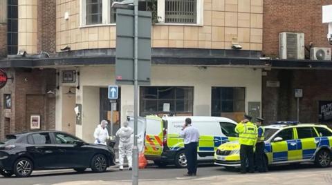 A liveried police car, a forensics van and a black car are parked nearby one another, with the rear doors of the van open and two people with white full-body suits stood by the rear of the van. There are also two uniformed police officers stood by the police car and a man in a white shirt and black trousers stood with his hand up to his ear.