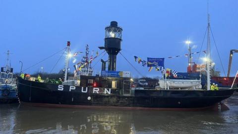 Spurn Lightship in Hull Marina. It  has a black hull with Spurn in white lettering in the centre is a black metal tower with the light at the top. There are ships flags and white lifeboats and people in high visibility jackets in the deck.