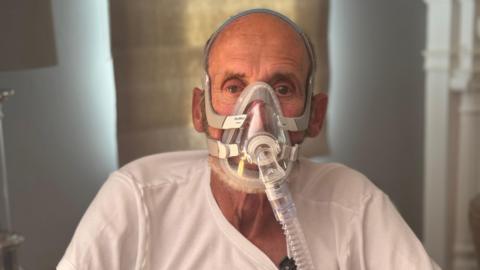 Paul stares straight to camera with a large plastic mask covering much of his face and tube hanging downwards. He wears a white T-Shirt.