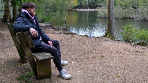 Connor Scott with short brown hair and short brown beard, wearing blue jacket and trousers with white trainers, sitting on a wooden bench overlooking a lake with trees.