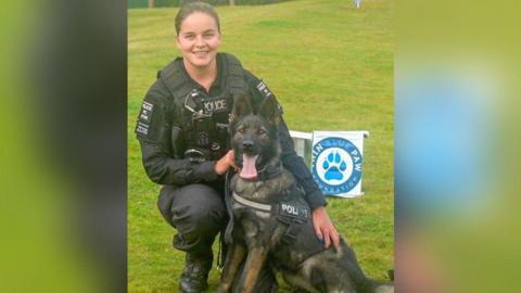 A female police officer smiles at the camera, squatting on the grass with her hands on a police dog. The dog is a German shepherd and has its tongue out, while wearing a strap that says police on it. A sign that says Thin Blue Paw foundation is visible in the background