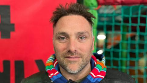 A man is standing in front of colourful soft play equipment. He is looking directly into the camera. He is wearing a colourful, striped hoodie underneath a black leather jacket.