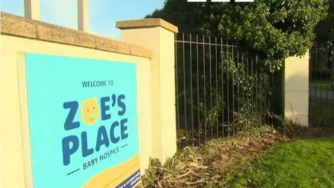 The Zoe's Place logo, which feature's blue letters and a teddy bear's face, on the yellow brick wall outside the hospice. 