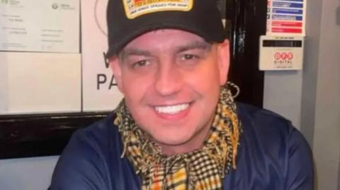 John George is smiling into the camera. He's wearing a black cap, with a design on it, and a navy top and a checked scarf.