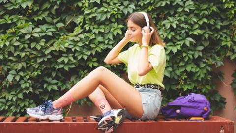 Teenager sitting on a bench in front of a green hedge. She is wearing a yellow shirt and putting headphones on over her ears.