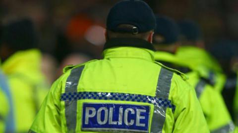 Police officers wearing high‑visibility yellow jackets with reflective stripes stand in a group, seen from behind, with “POLICE” printed on the back of the nearest jacket; several officers wear dark caps and carry helmets at waist level in a crowded outdoor setting.