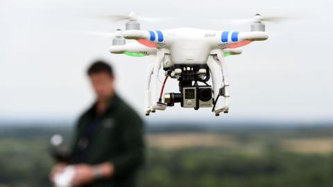 A drone in flight with the operator out of focus in the background.