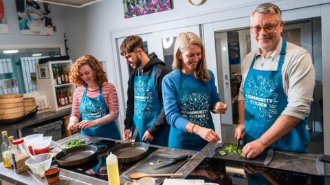 Two women and two men cooking using pans, saucepans and a chopping board.
