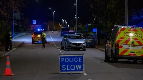 A police sign blocks a road at night illuminated by street lamps. An ambulance is at the right side of the road and a police officer can be seen next to a police car on the left. A slightly damaged car is in the centre of the road.