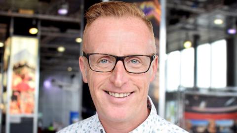 Bryan Burnett, a man with short orange hair which is slightly spiked up. He is wearing black glasses and a patterned shirt.