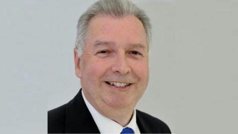 A professional headshot of a man with grey hair wearing a suit, smiling at the camera against a white background.