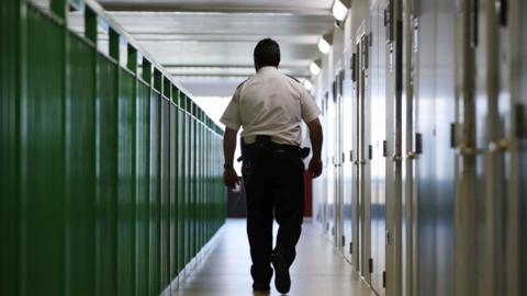 A prison guard walking through a corridor. Doors on the right, green fencing on the left.