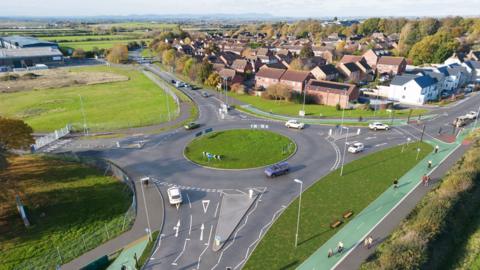 An artist's impression of the completed Bishop's Cleeve roundabout seen from above, with cars on the roundabout and houses also in the frame. Surrounding the road is a green two-lane cycle path.