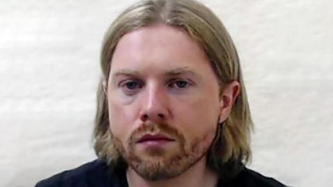 Mugshot of Luke Ford, who has chin-lenght blonde hair and a beard standing against a white wall wearing a black t-shirt.