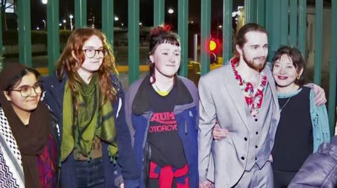Charlotte Head, Leona Kamio, Fatema Rajwani, Zoe Rogers and Jordan Devlin standing outside in the dark with a green, metal fence behind them. They are smiling.
