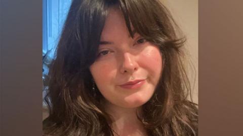 Chloe Humphries who has long dark wavy hair over her shoulders. Her head is tilted to her right and she has a serious expression on her face. 