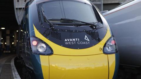 The mainly yellow and blue livery on the front of a train in a station. Avanti West Coast is written in white on black above the yellow.