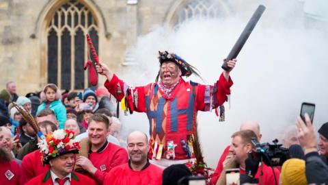 a man dressed in red with two letter H's on his top. holding a big pole and surrounded by smoke.